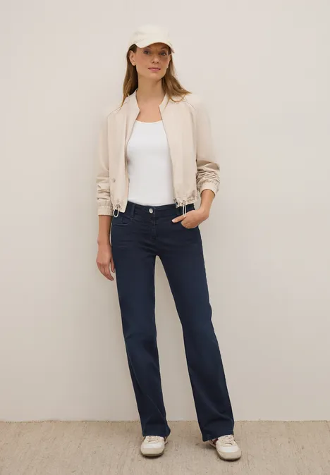 Frau mit heller Jacke, weißem Shirt, dunkler Hose und Cap steht entspannt.